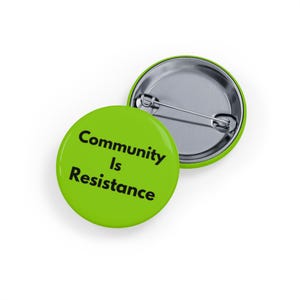 Puede incluir: Dos botones redondos de color verde lima con el texto "Community Is Resistance" en negro. Un botón está boca arriba y el otro boca abajo, mostrando el reverso plateado del alfiler.