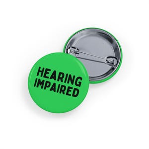 Könnte beinhalten: Zwei runde, grüne Ansteckbuttons mit dem schwarzen Schriftzug "HEARING IMPAIRED". Einer der Buttons liegt mit der Vorderseite nach oben, der andere mit der Rückseite, die die Nadel zeigt. Die Buttons dienen wahrscheinlich der Sensibilisierung oder Identifizierung.