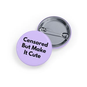 May include: Two round, light purple buttons. One button has the text "Censored But Make It Cute" in black. The other button is silver with a pin back. The buttons are on a white background.