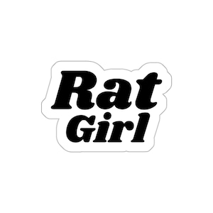 May include: A white sticker featuring the words "Rat Girl" in bold, black lettering with a retro aesthetic. The text is outlined in white, creating a layered effect. The sticker has a clean, minimalist design.
