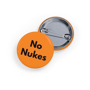 May include: Two orange round buttons with the text "No Nukes" in black. One button is face up, and the other is face down, showing the pin back. The buttons are likely for clothing or accessories.