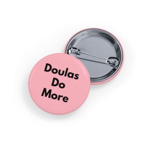 Puede incluir: Dos botones redondos rosas con el texto "Doulas Do More" en negro. Un botón está boca arriba y el otro boca abajo, revelando el alfiler. Los botones son de color rosa claro.