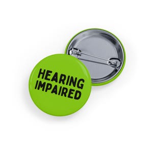 May include: Two round, lime green buttons with the words "HEARING IMPAIRED" in bold, black letters. One button is face up, and the other is face down, showing the silver pin back.