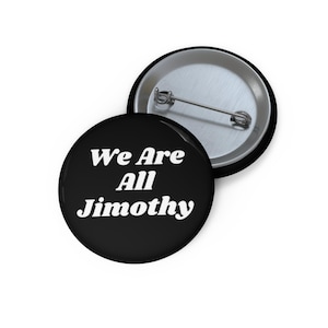 May include: A round black pin-back button featuring the white text "We Are All Jimothy" in a bold, sans-serif font. The back of the button is metallic silver and includes a standard horizontal pin mechanism for attachment to clothing or fabric items.