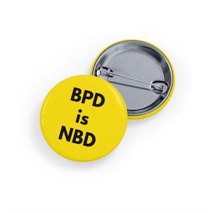 Może przedstawiać: Dwie żółte okrągłe przypinki z czarnym napisem "BPD is NBD". Jedna przypinka jest skierowana przodem do góry, a druga tyłem do dołu, pokazując agrafkę. Przypinki są na białym tle.