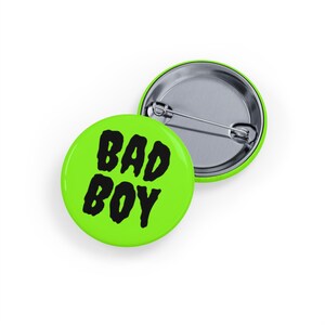 May include: Two round, lime green pin badges. One badge displays the words "BAD BOY" in bold, black letters. The second badge is silver with a safety pin backing. The badges are on a white background.