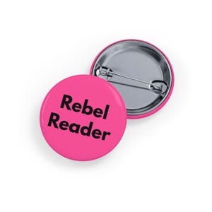 Könnte beinhalten: Zwei runde, leuchtend pinkfarbene Ansteckbuttons mit silberner Nadelrückseite. Der vordere Button zeigt die Worte "Rebel Reader" in fetter, schwarzer Schrift. Die Buttons sind für Kleidung oder Accessoires geeignet.