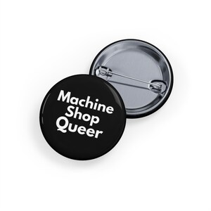May include: Two round black pinback buttons. One button displays the text "Machine Shop Queer" in white. The other button is silver with a pin. The buttons are on a white background.