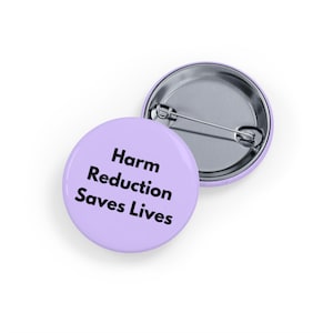 Puede incluir: Dos insignias redondas de color lila claro con respaldos de alfiler plateados. Una insignia muestra el texto "Harm Reduction Saves Lives" en negro y en negrita. La segunda insignia es parcialmente visible, mostrando el respaldo del alfiler plateado.