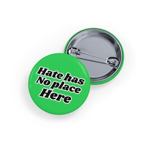 May include: Two green round buttons with the text "Hate has No place Here" in black and white. One button is face up, and the other is face down, showing the pin back.