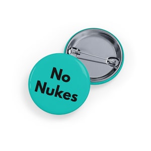May include: Two round teal buttons with the words "No Nukes" in black. One button is face up, and the other is slightly angled, revealing the pin back. The buttons are likely made of metal and plastic.