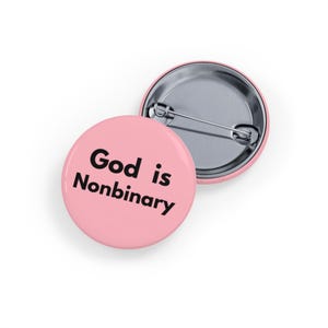 Puede incluir: Dos insignias redondas rosas. Una insignia muestra el texto "God is Nonbinary" en negro, con fuente sans-serif. La otra insignia es plateada y tiene un alfiler en la parte posterior.