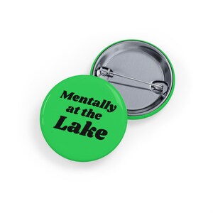 Peut inclure: Deux badges ronds verts. L'un porte le texte "Mentally at the Lake" en noir. L'autre est argenté avec un bord vert. Les badges sont sur fond blanc.