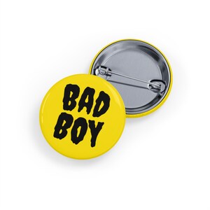 May include: Two yellow round pin badges. One badge has the words "BAD BOY" in bold black letters. The other badge is silver on the back. The badges are on a white surface.