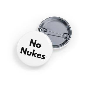 May include: Two white round buttons with the text "No Nukes" in bold black letters. One button is face up, and the other is face down, revealing the pin back. The buttons are on a white background.