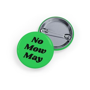 May include: Two green round buttons with the text "No Mow May" in black. One button is face up, and the other is face down, showing the pin back. The buttons are on a white background.