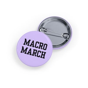 Peut inclure: Deux badges ronds, violets clairs, avec le texte "MACRO MARCH" en lettres noires en gras. Un badge est face visible et l'autre est face cachée, révélant l'épingle.