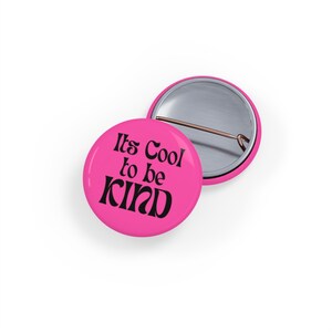 Può includere: Bottone rosa con il testo "It's Cool to be Kind" in lettere nere.