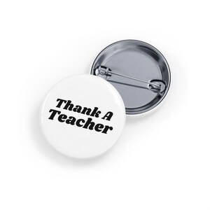 Könnte beinhalten: Zwei weiße, runde Ansteckbuttons mit Nadelverschluss. Ein Button zeigt den Text "Thank A Teacher" in schwarzer, fetter Schrift. Der andere Button ist teilweise sichtbar und zeigt die Nadel. Die Buttons befinden sich auf weißem Hintergrund.
