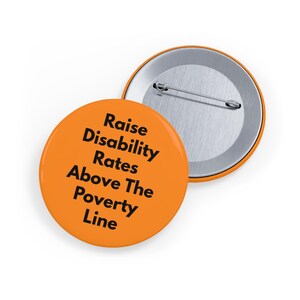 May include: Two orange round badges with the text "Raise Disability Rates Above The Poverty Line" in black. One badge is in focus, the other is partially visible. The badges have a silver pin backing.