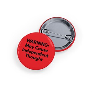 Può includere: Due bottoni rotondi rossi con spilla di sicurezza. Uno mostra il testo nero "WARNING: May Cause Independent Thought". L'altro bottone mostra il retro e la spilla.