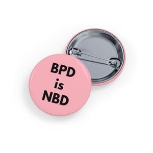 Może przedstawiać: Dwa okrągłe, różowe guziki z metalowym tyłem na szpilkę. Jeden guzik wyświetla tekst "BPD is NBD" czarnymi, pogrubionymi literami. Drugi guzik jest częściowo widoczny, pokazując metalowy tył. Guziki leżą na białej powierzchni.