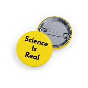 May include: Two yellow, round badges with the text "Science Is Real" in black. One badge is face up, and the other is face down, showing the pin back. The badges are on a white background.