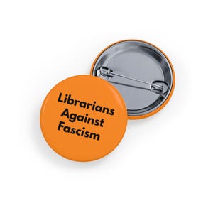 Op de afbeelding: Twee oranje ronde buttons met een zilveren speldrug. De voorste button heeft de tekst "Librarians Against Fascism" in zwart, gecentreerd op de button.