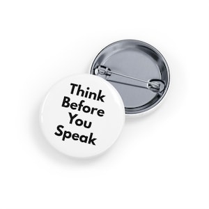 May include: Two white round badges with the text "Think Before You Speak" in black. One badge is face up, and the other is face down, showing the pin back. The badges are on a white surface.