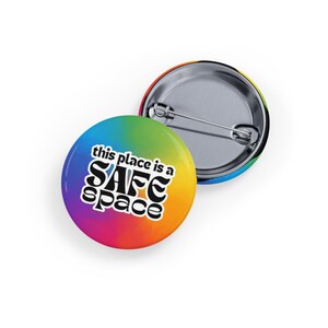 Pode incluir: Um botão de cor arco-íris com o texto "this place is a safe space" em letras pretas. O botão é circular e tem um verso de metal.