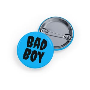 May include: Two round badges. One is light blue with the words "BAD BOY" in black. The other badge is light blue with a silver pin back. The badges are on a white background.