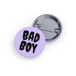 May include: Two round, pale purple badges with the words "BAD BOY" in bold, black letters. One badge is face up, and the other is face down, showing the pin back.