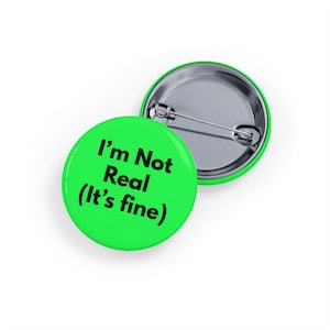 May include: Two round, green pin badges. One badge displays the text "I'm Not Real (It's fine)" in black. The other badge is partially visible, showing the silver pin backing and a green border. These badges are likely for clothing or accessories.
