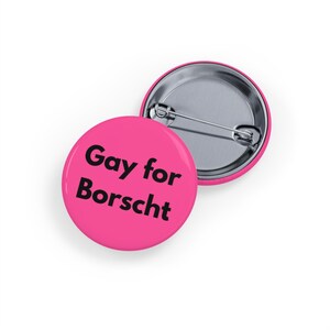 Peut inclure: Deux badges ronds roses. L'un affiche le texte "Gay for Borscht" en lettres noires et grasses. L'autre badge est partiellement visible, montrant le dos de l'épingle argentée. Les badges sont sur fond blanc.