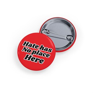 May include: Two red round buttons with a silver pin backing. The front button has the text "Hate has No place Here" in white, outlined in black. The second button is partially visible.
