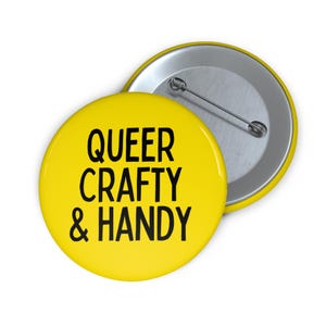 May include: A yellow button with the text "QUEER CRAFTY & HANDY" in black. The button has a silver pin back. The button is round and has a glossy finish. The button is a craft supply.