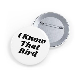May include: A round pin-back badge featuring a white surface with the black text I Know That Bird printed in a bold serif font. The reverse of the badge is metallic silver with a horizontal pin mechanism for attachment to clothing or fabric items.