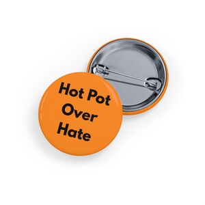 May include: Two orange round badges with a safety pin fastening. The front badge has the words "Hot Pot Over Hate" in black text. The second badge is partially visible, showing the silver metal backing.