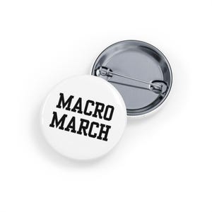 May include: Two white, round badges with the text "MACRO MARCH" in bold black letters. One badge is face up, and the other is face down, showing the pin back. The badges are on a white background.
