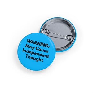 Può includere: Due bottoni rotondi azzurri con retro a spillo argentato. Un bottone è rivolto verso l'alto, l'altro è leggermente angolato. Il testo "WARNING: May Cause Independent Thought" è stampato in nero sulla parte anteriore del bottone.