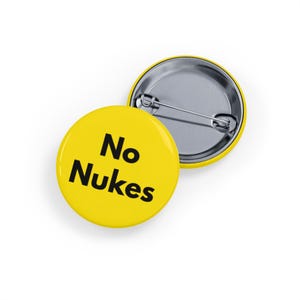 May include: Two yellow round buttons with the text "No Nukes" in black. One button is face up, and the other is face down, showing the pin back. The buttons are on a white surface.