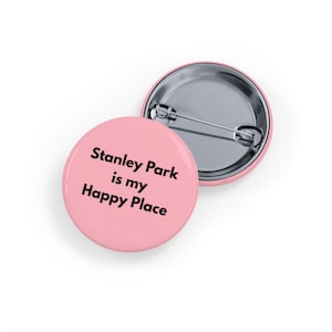 May include: Two round, pink pinback buttons. One button displays the text "Stanley Park is my Happy Place" in black. The other button is silver. The buttons are on a white background.