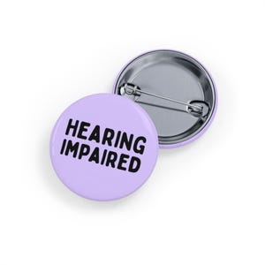 Puede incluir: Dos insignias redondas de color lila claro con la inscripción "HEARING IMPAIRED" en letras negras en negrita. Una insignia está delante de la otra, mostrando la parte trasera del alfiler. Estas insignias son probablemente para la concienciación o la identificación.