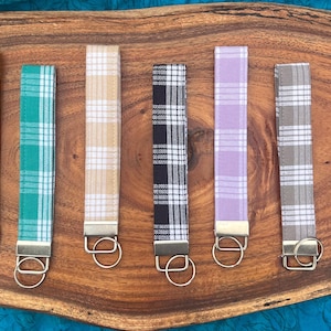 May include: Eight keychains with different plaid patterns. The keychains are made of fabric and have a silver metal ring. The keychains are arranged in a row on a wooden surface.