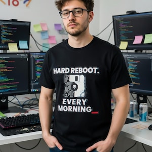 May include: Black t-shirt with the text "HARD REBOOT. EVERY MORNING" in white. The graphic includes a floppy disk, skull, and circuit board design. The person is wearing glasses and standing in front of computer monitors.
