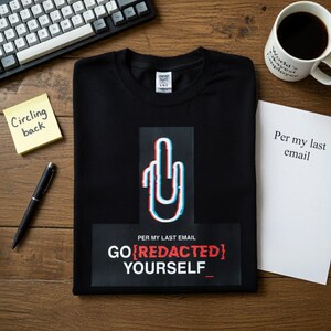 Op de afbeelding: Zwart T-shirt met een afbeelding van een handgebaar gemaakt van een paperclip met rode en blauwe accenten. De tekst "GO REDACTED YOURSELF" staat eronder. Een koffiemok, toetsenbord, pen en plakbriefje zijn ook zichtbaar.