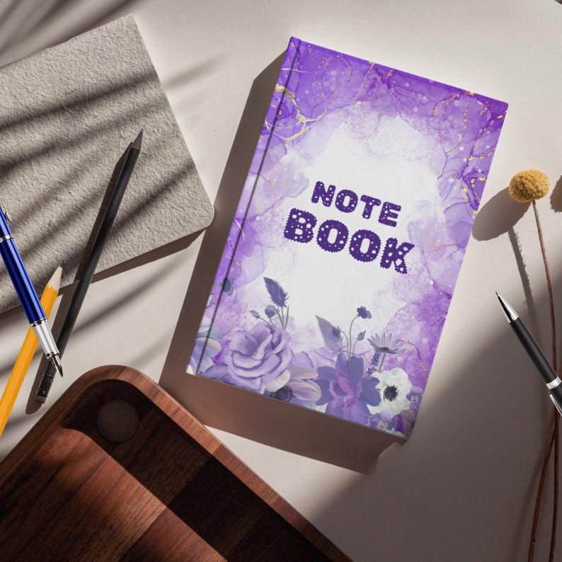 Puede incluir: Un cuaderno morado con la palabra "NOTE BOOK" en fuente punteada. La portada presenta un dise&ntilde;o de acuarela con tonos morados, detalles dorados e ilustraciones florales. Un bol&iacute;grafo, un l&aacute;piz y otros art&iacute;culos de papeler&iacute;a est&aacute;n colocados alrededor.