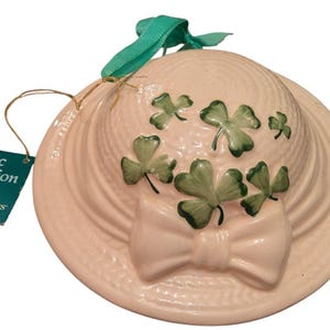 Puede incluir: Un sombrero de cerámica blanco con un diseño de trébol verde. El sombrero tiene una cinta verde y está decorado con un lazo blanco. El sombrero forma parte de la colección Celtic de Triple Dreams.
