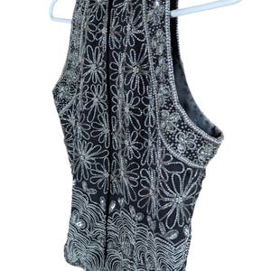 May include: Black sleeveless top with intricate silver bead and sequin floral and geometric patterns. The top is hanging on a white hanger. The embellishments create a shimmering effect, perfect for special occasions.
