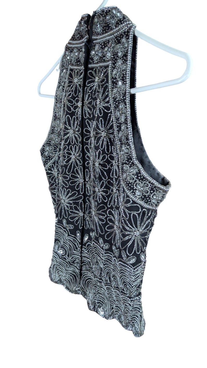 May include: Black sleeveless top with intricate silver bead and sequin floral and geometric patterns. The top is hanging on a white hanger. The embellishments create a shimmering effect, perfect for special occasions.